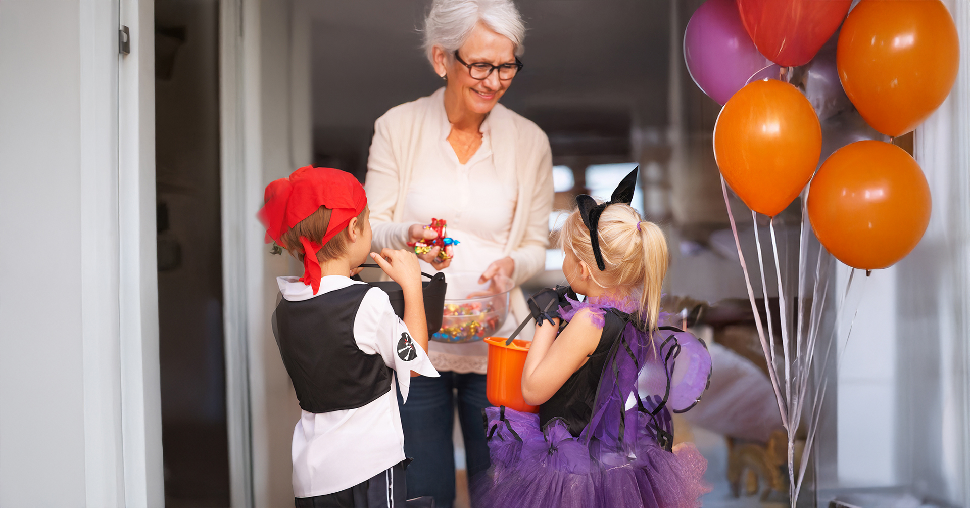 A senior woman handing out candy to 2 children dressed up for Halloween Trick-or-Treating